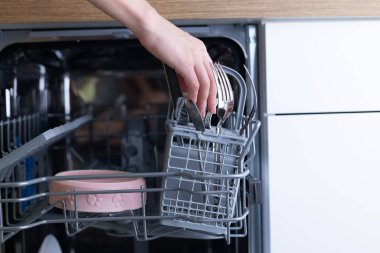Bu resim, çatalları bulaşık makinesine yerleştiren bir el görüntüsü yakalıyor, modern mutfak aletlerinin kolaylığını ve pratik bulaşık yıkama çözümlerini vurguluyor..