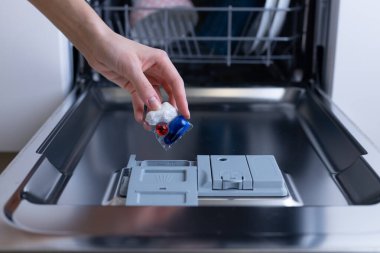 Bu resim, modern bulaşık sistemlerinin kolaylığını ve etkinliğini vurgulayarak, bir bulaşık makinesinin belirlenmiş kompartımanına renkli bir deterjan kapsülü koyan bir insanı gösteriyor..