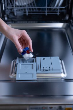 Bulaşık makinesine renkli bir deterjan koyan, modern mutfak hijyenini vurgulayan ve bulaşık malzemeleri için temizlik teknolojisini kullanan bir kişinin yakın çekim görüntüsü..