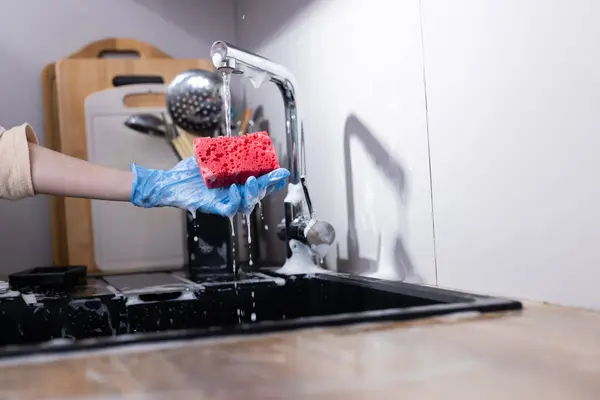 Görüntü, elinde parlak kırmızı bir sünger tutan mavi eldiven takan birini gösteriyor. Her yerde sabun köpükleri olan modern bir mutfakta musluktan su akıyor..