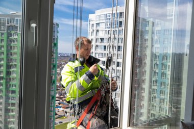 Parlak güvenlik ceketli bir işçi, yüksek bir yerden askıdayken pencere temizliği yapıyor. Gökdelenler, canlı mavi gökyüzü ile çevrili, kentsel yaşam ve bakım çalışmaları sergiliyorlar..