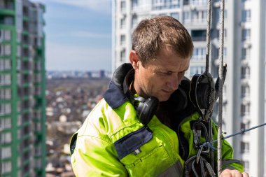 Parlak güvenlik teçhizatı içinde yetenekli bir işçi, yoğun şehir ortamında yüksek bir binada asılı dururken titizlikle ekipmanları hazırlıyor..