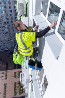 Görüntü, parlak sarı bir gömlekle bir pencere temizleyicisini yakalıyor. Yüksek binadaki camları ustalıkla temizliyor. Altında canlı bir şehir manzarası var..