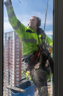 Görüntü, kendini adamış parlak sarı ceketli bir pencere temizleyicisinin yer üstünde çalışarak mavi gökyüzü altında hareketli bir şehir ortamında beceri ve hassasiyet gösterdiğini gösteriyor..