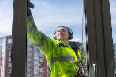 Çalışkan bir pencere temizleyicisinin, yüksek bir binanın camını ovaladığı, parlak sarı bir güvenlik önlüğü ve kulak koruyucusu giydiği, güvenlik ve temizliğe olan bağlılığını gösterdiği görülüyor..