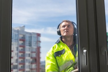 Görüntü, sarı bir ceketle yukarı bakan ve kulaklık takan bir cam temizleyicisini gösteriyor. Şehir manzarası ve mavi gökyüzü arka planda görünürken o dışarıda çalışıyor..