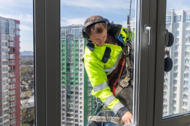 Parlak güvenlik kıyafeti ve kulaklıkla donatılmış özel bir pencere temizleyicisi, yüksek katlı pencereleri temizliyor ve zorlu işlerde güvenlik ve profesyonelliğin önemini gösteriyor..