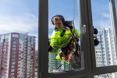 Yüksek katlı bir binanın önünde camları titizlikle temizleyen yetenekli bir pencere temizleyicisi asılı duruyor. Sahnede kent mimarisi, güvenlik ekipmanları ve arkaplan olarak parlak mavi bir gökyüzü sergileniyor..
