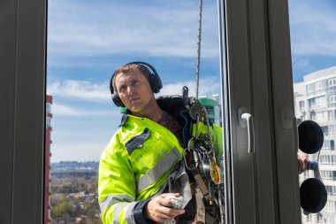 Kulaklıklar ve emniyet kemerleriyle donatılmış, parlak güvenlik teçhizatı olan bir pencere temizleyicisi, yüksek katlı pencereleri temizleme görevine odaklanıyor. Şehir silueti ve mavi gökyüzü çarpıcı bir zemin oluşturuyor..