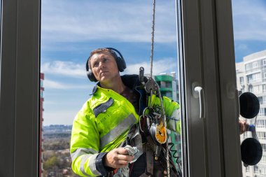 Parlak sarı bir güvenlik ceketi giymiş kendini adamış bir işçi, yüksek katlı bir binanın camlarını temizliyor, şehir bakımını ve inşaat alanındaki güvenlik teçhizatının önemini gösteriyor..