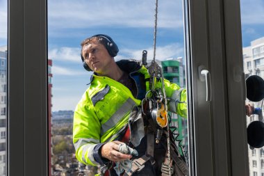 Yüksek bir binanın önünde asılı duran, parlak güvenlik ekipmanları ve kulaklıklar takan, görev sırasında güvenliği sağlarken profesyonelliği gösteren özel bir pencere temizleyicisi.