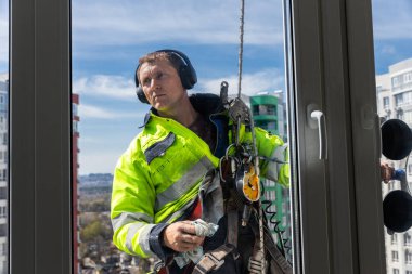 Yüksek görüşlü ceketli ve güvenlik teçhizatlı bir cam temizleyicisi yüksek katlı bir bina üzerinde çalışıyor. Kulaklık takıyor ve arkasında güzel bir şehir manzarası var..