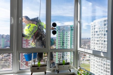 Görüntü, şehir manzarasının panoramik manzarası ve altındaki yeşillik ile yüksek camlarda çalışan parlak sarı ceketli profesyonel bir pencere temizleyicisini yakalıyor..