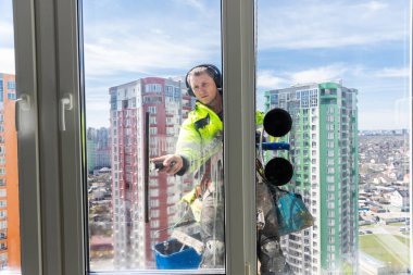 Görüntü, arka planda parlak mavi gökyüzü ve modern şehir mimarisi ile yüksek bir binanın camlarını ustalıkla silen, güvenlik donanımlı yetenekli bir pencere temizleyicisini gösteriyor..