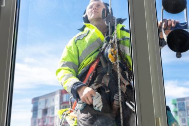 Parlak bir güvenlik ceketi giymiş odaklanmış bir cam temizleyicisi, özenle yüksek bir mevkide çalışıyor. Aletleri ve koşumları riskli işlerde güvenliğin önemini vurguluyor..