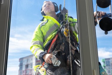 Görüntü, yüksek görüş açısına sahip bir ceketle kendini adamış bir tamirciyi, yüksek bir binanın dış yüzeyinde güneşli bir zemine karşı çalışırken güvenlik ekipmanları kullanarak resmediyor..