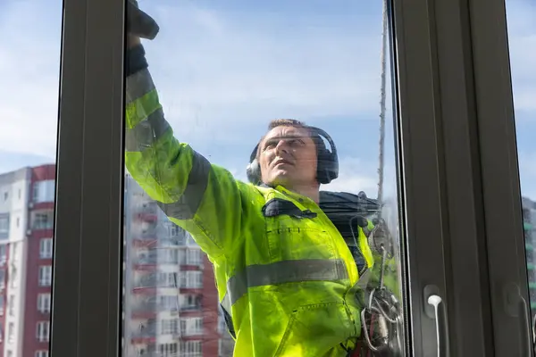 Çalışkan bir pencere temizleyicisinin, yüksek bir binanın camını ovaladığı, parlak sarı bir güvenlik önlüğü ve kulak koruyucusu giydiği, güvenlik ve temizliğe olan bağlılığını gösterdiği görülüyor..