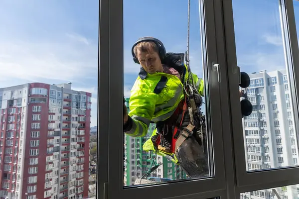 Yüksek katlı bir binanın önünde camları titizlikle temizleyen yetenekli bir pencere temizleyicisi asılı duruyor. Sahnede kent mimarisi, güvenlik ekipmanları ve arkaplan olarak parlak mavi bir gökyüzü sergileniyor..