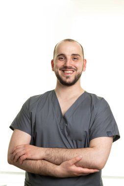 Gri önlük giyen neşeli bir erkek sağlık çalışanı kollarını kavuşturmuş, minimalist bir ortamda dostça bir gülümseme sergiliyor..