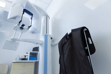 Klinik bir ortamda sergilenen parlak bir diş görüntüleme makinesi. Yakınlarda asılı koruyucu bir kurşun önlük var. Radyografik işlemler sırasında hastanın güvenliğini sağlıyor..