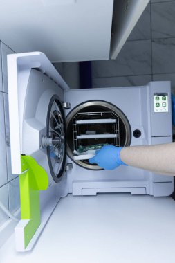 Yakından çekilmiş bir görüntü sterilizasyon cihazını gösteriyor. Eldivenli bir el dezenfekte için bir şeyler yerleştiriyor. Ekipman, tıbbi hazırlık uygulamalarında verimliliği vurguluyor.