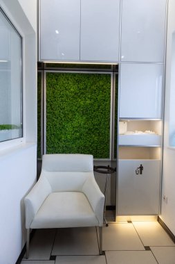 Lüks beyaz bir sandalye ve canlı yeşil bir yosun duvarının kombinasyonunu gösteren şık bir bekleme salonu, modern estetiği güçlendirirken aynı zamanda rahatlatıcı ve davetkar bir atmosfer sağlıyor..