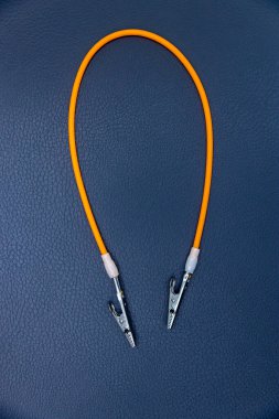 Görüntü her iki ucunda iki timsah klipsi bulunan turuncu bir elektrik kablosu sergiliyor, koyu mavi bir arkaplana yaslanmış. Bu ayar elektrik işlerinde kullanışlı olduğunu gösteriyor.