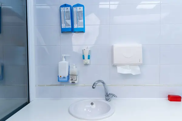 Görüntü, sabun makineleri, kağıt havlu tutacağı ve beyaz fayanslı duvarlara karşı parlak bir banyo lavabosu ve çeşitli hijyen malzemelerini yakalıyor..