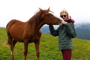 Büyüleyici bir sahne, genç bir kadının canlı yeşil bir alanda çarpıcı bir atla yakından etkileşimini gösteriyor. Dağlarla çevrili doğanın güzelliğini yansıtıyor..
