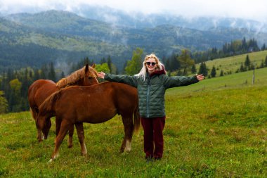Bir kadın açık kollarla ayakta duruyor, canlı yeşil bir çayırda iki atla neşe içinde etkileşime geçiyor, görkemli dağlarla çevrili, parçalı bulutlu bir gökyüzünün altında, doğanın güzelliğini gösteriyor..