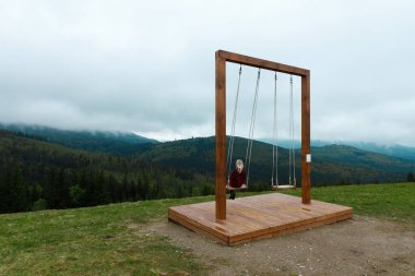 Görüntü, bulutlar ve yemyeşil yemyeşil gölgelerle örtülmüş görkemli dağların zemininde huzurlu bir ahşap salıncak sergiliyor, doğanın kucaklamasında rahatlamak için mükemmel..