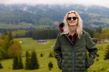 Görüntü, şık bir ceket ve güneş gözlüğü giymiş, yemyeşil bir dağlık alanda duran neşeli bir kadını yansıtıyor. Resimli bir arkaplan huzurlu atmosferi güçlendirir.