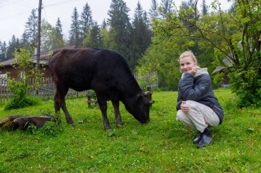 Huzurlu bir kırsal alanda, genç bir kadın otlayan bir ineğin yanında çömelmiş, yeşillik, ağaçlar ve arka planda kırsal bir cazibe ile çevrili, çiftlik hayatını gözler önüne seriyor..