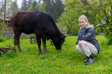 Siyah bir ineğin yanında çömelmiş, arka planda ağaçlar ve geleneksel ahşap binalarla çiftliğin taze yeşilliğinin tadını çıkaran genç bir kadının yer aldığı huzurlu bir sahne..