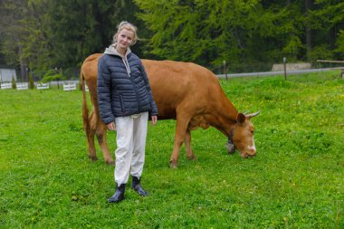 Gündelik giysiler giymiş genç bir birey yemyeşil bir alanda otlayan kahverengi bir ineğin yanında duruyor. Bu da çiftçilik ortamında insanlarla doğa arasında huzurlu bir bağ olduğunu gösteriyor..