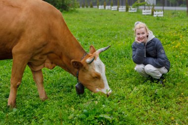 Sakin bir kırsal alanda, bir kadın taze çimenlerde mutlu bir şekilde otlayan kahverengi bir ineğin yanında çömelir, canlı yeşillikler arasında insanlarla hayvanlar arasında güzel bir bağ olduğunu gösterir..