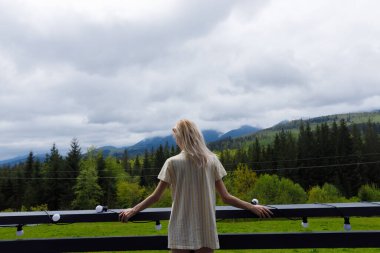 Bu sakin sahnede, genç bir kadın balkonda duruyor, nefes kesen dağ manzarasına bakıyor, bulutlu bir günde, yemyeşil bir hava onu çevrelemiş..