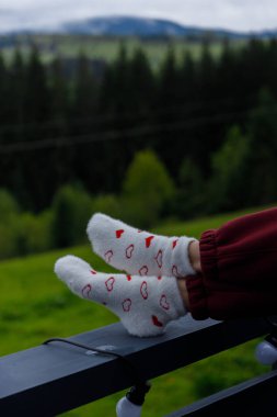Kırmızı kalp desenleriyle süslenmiş şık beyaz çoraplar balkonda rahatça dinleniyor. Manzaralı arka planda yeşil tepeler ve sakin bir orman manzarası var..