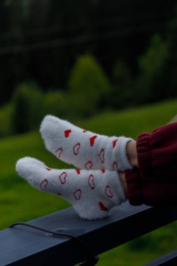 Kırmızı kalp desenleriyle bezenmiş bir çift tüylü beyaz çorap balkonda rahatça dinleniyor arka planda yemyeşil bir korkuluk, sıcaklığı ve rahatlığı simgeliyor.. 