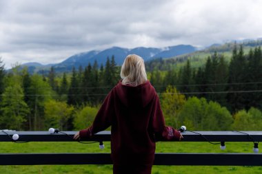 Bu büyüleyici sahnede, bir kadın balkonda duruyor nefes kesici dağlara ve bol yeşilliğe bakıyor, sükuneti somutlaştırıyor ve manzaranın doğasıyla bağlantı kuruyor..