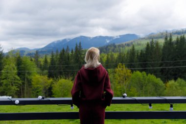 Huzurlu görüntü, verandasındaki güzel dağlara ve ağaçlara bakarak doğanın cazibesiyle huzurlu ve ilham verici bir atmosfer yaratan bordo kıyafetli bir kadını yakalıyor..