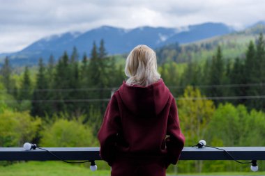 Bu sakin sahnede, bir kadın balkonda duruyor, yükselen dağlara ve yoğun yeşil ormanlara bakıyor, doğayla derin bir bağ kuruyor ve günlük kıyafetlerini giyiyor..