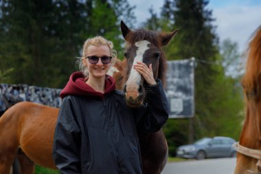 Bu güzel sahnede, gülümseyen genç bir kadın bir atla nazikçe iletişim kurar, insanlar ve hayvanlar arasındaki bağı gözler önüne serer, çarpıcı bir doğa zeminine karşı kurulur..