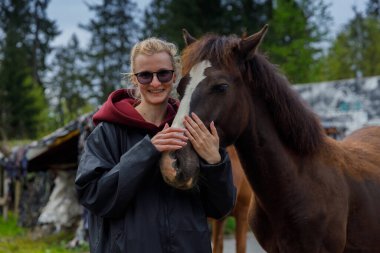 Bu iç açıcı sahnede, gülümseyen genç bir kadın narin bir atla poz veriyor, bağlarını sakin doğal bir arka planda sergiliyor ve vahşi yaşama karşı derin bir takdiri yansıtıyor..