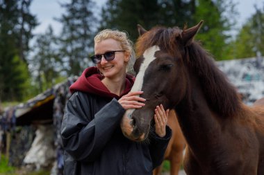 Bu iç açıcı sahnede, gülümseyen genç bir kadın bir atla sevgi dolu bir şekilde etkileşime geçiyor, insanlar ve hayvanlar arasındaki bağın huzurlu ve doğa dolu bir ortamda sergilendiğini gösteriyor..