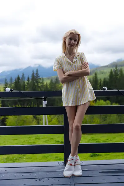 Görüntüde, sarı çizgili sıradan bir kıyafet giymiş, balkonda duran genç bir kadın görülüyor. Arkaplan bulutlu bir gökyüzünün altında, sükuneti çağrıştıran güzel yeşil dağları vurguluyor..