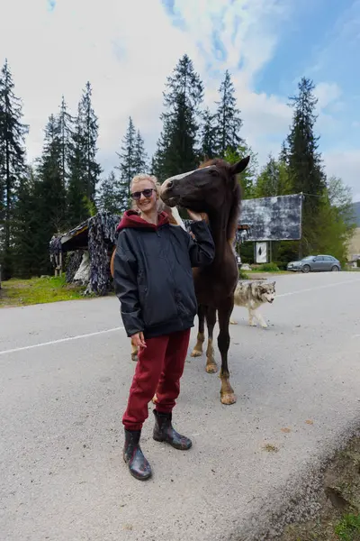 Bu huzurlu manzarada, mutlu bir insan bir atla nazikçe etkileşime geçerken gülümser, yemyeşil bir ortamda ve yakınlardaki meraklı bir köpeğin arasında hoş bir bağlantı gösterir..