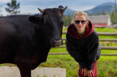 Görüntü, güneş gözlüğü takan genç bir kadını ve siyah ceketini canlı yeşil tarlalar ve dağlarla çevrili bir çiftlikte, parçalı bulutlu bir gökyüzünün altında siyah bir ineğin yanında poz verirken yakalıyor..