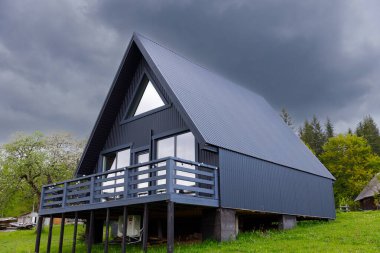 Bu görüntü, parlak, koyu renkli dış görünüşlü, geniş pencereli, doğal ışığı yakalayan şık bir A-çerçeve ev ve güzel yeşil ortamda rahatlamak için mükemmel bir güverte içerir..