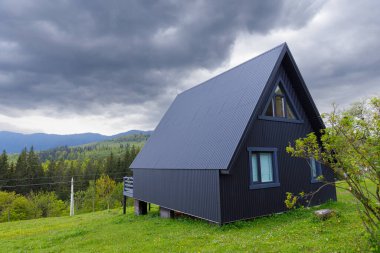 Pürüzsüz, siyah bir çerçeve ev, canlı yeşil ve dramatik bulutlarla çevrili bir yamaçta gururla durur. Modern tasarımın doğanın güzelliğiyle uyumunu gözler önüne serer..
